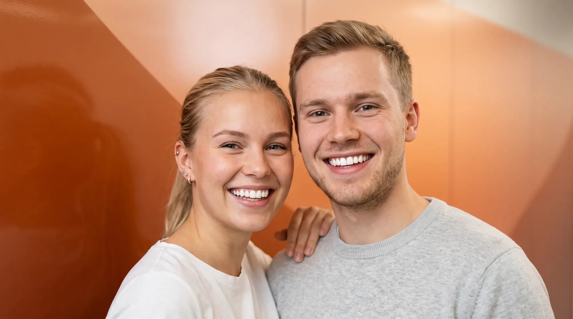 Ung mann og kvinne smiler med hvite tenner mot oransje gradientbakgrunn